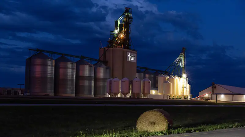 Un terminal céréalier intérieur illuminé et des cellules en acier au crépuscule en Saskatchewan, une balle ronde dans le fossé