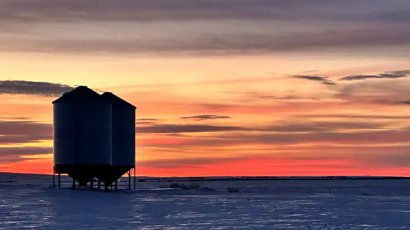A steel grain bin on a snowy Saskatchewan field under an orange winter sunset